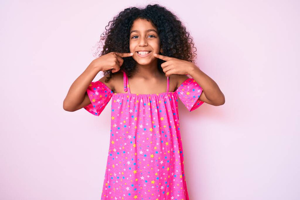 Africké americké dítě s kudrnatými vlasy na sobě ležérní šaty s veselým úsměvem ukazující a ukazující s prsty zuby a ústa. koncepce stomatologického zdraví.  - Fotografie, Obrázek