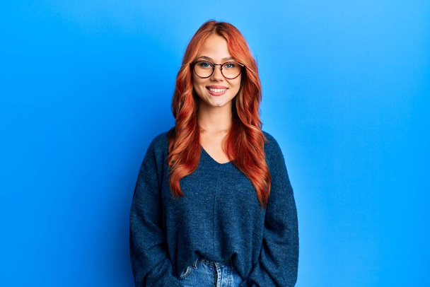 Young beautiful redhead woman wearing casual sweater and glasses over blue background with a happy and cool smile on face. lucky person.  - Photo, Image