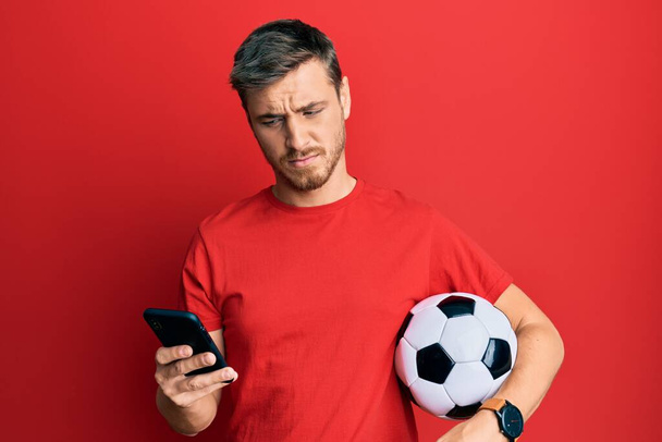 Όμορφος καυκάσιος άντρας που κρατάει μπάλα ποδοσφαίρου κοιτάζοντας το smartphone στο πρόσωπο, δείχνοντας σκεπτικός και σαρκαστικός, έκπληκτος με ανοιχτό στόμα  - Φωτογραφία, εικόνα