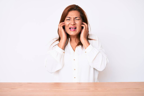 Femme d'affaires hispanique brune du moyen âge portant une chemise blanche décontractée assise sur la table couvrant les oreilles avec des doigts avec une expression agacée pour le bruit de la musique forte. concept sourd.  - Photo, image
