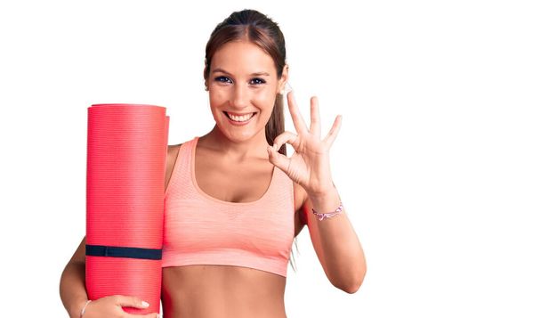 Young beautiful hispanic woman holding yoga mat doing ok sign with fingers, smiling friendly gesturing excellent symbol  - Φωτογραφία, εικόνα