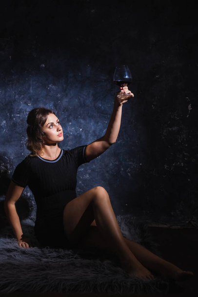 Beautiful young lady in black dress sitting on fur skin with glass of wine - Zdjęcie, obraz
