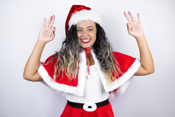 Young beautiful woman wearing a Santa Claus costume over white background doing ok sign with fingers and smiling, excellent symbol - Foto, Imagem