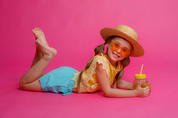 uma menina em óculos de sol, um chapéu de palha e um coquetel está deitado contra o fundo rosa  - Foto, Imagem