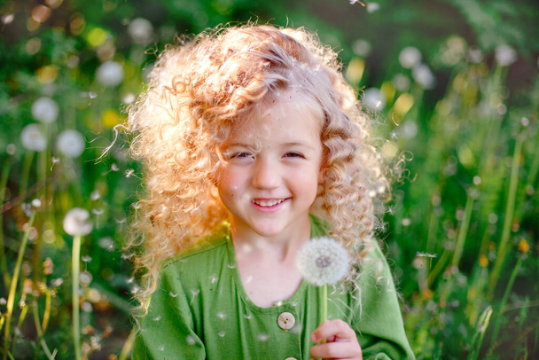 kleine lockige blonde Mädchen in grünem Kleid weht halten Löwenzahn im Park Lächeln  - Foto, Bild