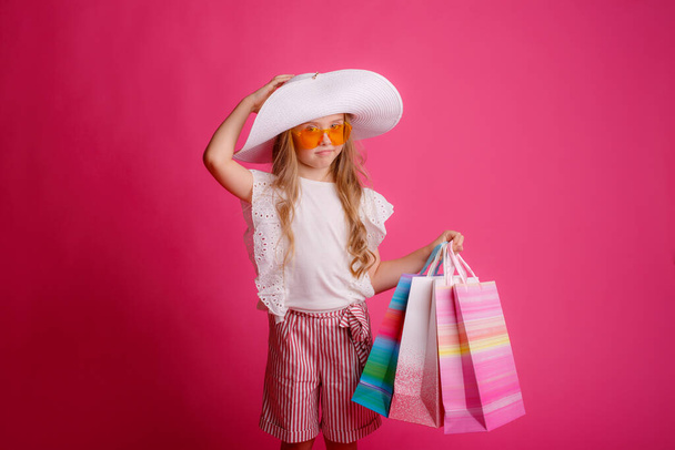 una niña con un montón de bolsas de compras, con gafas de sol y un sombrero sobre un fondo rosa en el estudio - Foto, Imagen