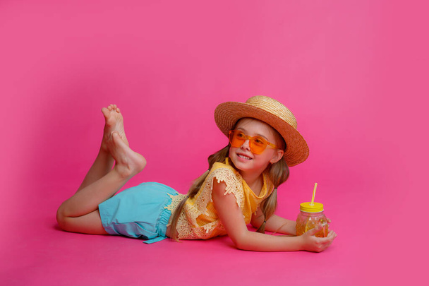 une petite fille en lunettes de soleil, un chapeau de paille et un cocktail est couché sur fond rose  - Photo, image