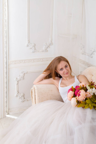 Beautiful young woman with flowers dressed in a white full wide-skirted dress sitting on the sofa - Foto, Imagen