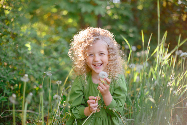 Küçük kıvırcık saçlı, yeşil elbiseli kız. Parktaki tebessümde karahindiba esiyor.  - Fotoğraf, Görsel