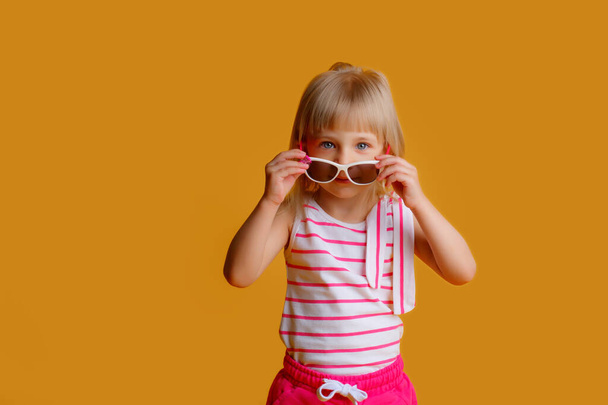 een klein blond meisje in zomerkleren en een zonnebril op een gele achtergrond. Het concept van de zomervakantie - Foto, afbeelding