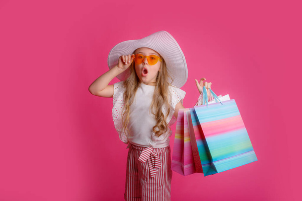 a little Girl with a lot of shopping bags, wearing sunglasses and a hat on a pink background in the Studio - Foto, imagen