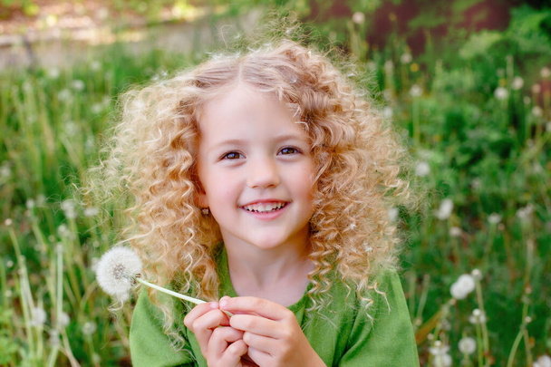 小さな巻き毛のブロンド女の子で緑のドレス吹いてホールドタンポポで公園の笑顔  - 写真・画像