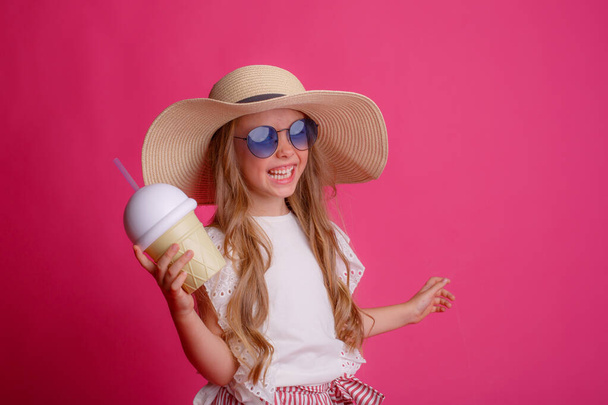 πορτρέτο μιας μικρής ξανθιάς με γυαλιά ηλίου και ένα καπέλο σε ροζ φόντο με μια κονσέρβα δροσιστικό κοκτέιλ. - Φωτογραφία, εικόνα