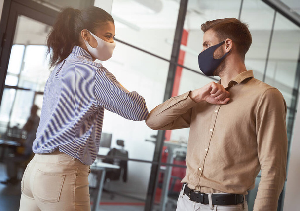 Dos jóvenes colegas de negocios diversos que usan máscaras protectoras de la cara golpeando codos, saludándose mientras trabajan durante la cuarentena covid 19 - Foto, imagen