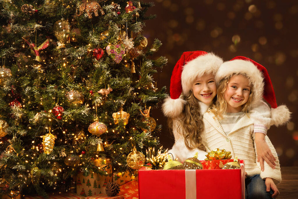 Christmas Children Sisters opening Present Box with Gift Toys. Happy smiling little Girls in Santa Hat near to Christmas tree in dark background - Foto, Imagem