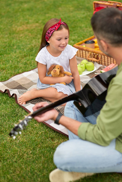 Αξιολάτρευτο κοριτσάκι που κάθεται σε ένα πράσινο γρασίδι στο πάρκο και κοιτάζει τον πατέρα της να παίζει κιθάρα. - Φωτογραφία, εικόνα