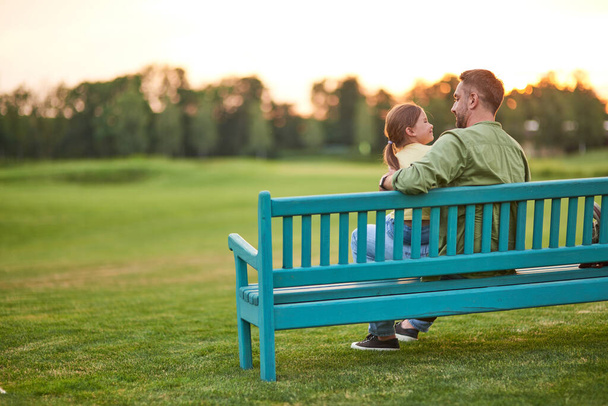 Dlouhý záběr, jak mladý otec tráví čas se svou roztomilou dceruškou. Dívají se na sebe, zatímco sedí v létě na lavičce v parku. - Fotografie, Obrázek