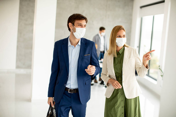 Bello giovane coppia d'affari a piedi insieme mentre indossano maschere protettive per evitare corona viroso in ufficio - Foto, immagini