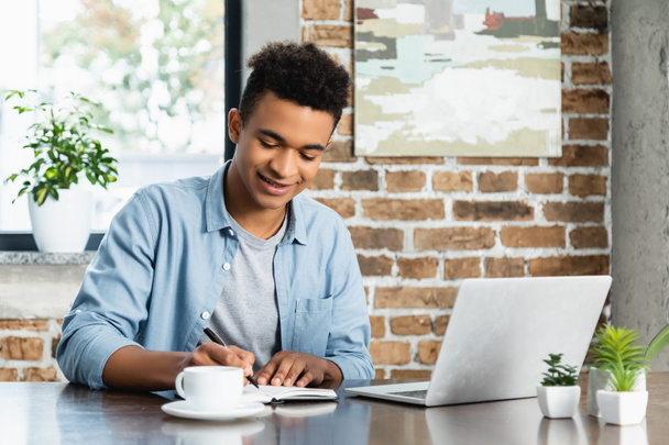 hombre afroamericano feliz escribiendo en un cuaderno cerca de la computadora portátil y la taza en el escritorio - Foto, imagen