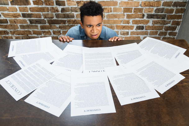 шокированный африканский американец, рассматривающий документы с контрактными надписями на столе - Фото, изображение