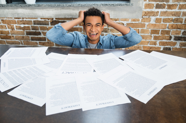 histérico afroamericano hombre tirando de pelo cerca de documentos con letras de contrato en el escritorio - Foto, imagen