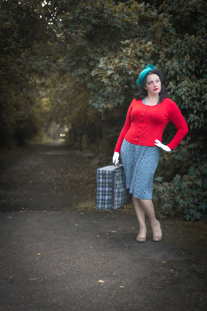 Beautiful young woman posing in vintage 1940s clothes, waiting by trees with suitcase - Foto, Imagem