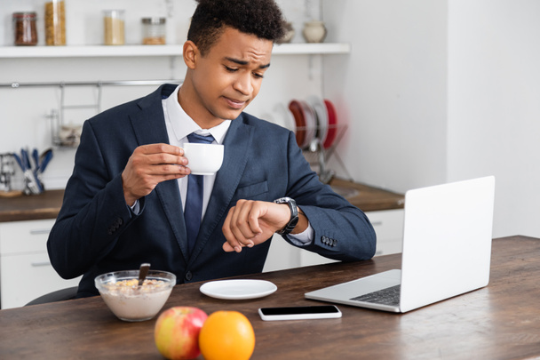 afrikaanse amerikaanse freelancer in pak holding cup en kijken naar horloge in de buurt gadgets op bureau - Foto, afbeelding