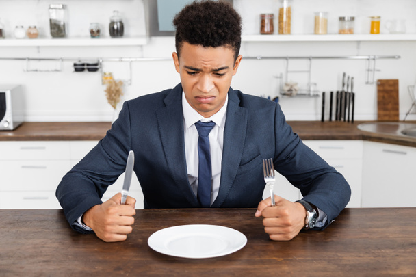frustrated african american man holding cutlery and looking at empty plate - Фото, изображение