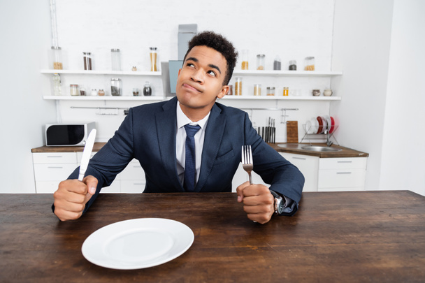 dreamy african american man holding cutlery while thinking and sticking out tongue  - Photo, Image