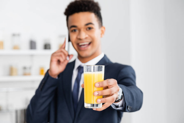 glass of orange juice in hand of happy african american businessman talking on smartphone on blurred background  - Foto, immagini