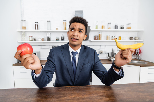 μπερδεμένος Αφροαμερικανός επιχειρηματίας που επιλέγει μεταξύ μήλου και μπανάνας - Φωτογραφία, εικόνα
