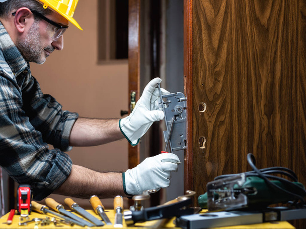 Een close-up. Timmerman op het werk repareert en installeert het nieuwe slot van een houten deur. Bouwsector.  - Foto, afbeelding