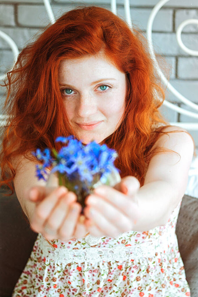 Söpö punapää tyttö istuu suuressa tuolissa, jossa on kukkakimppu luonnonvaraisia sinisiä lumikelloja käsissään.. - Valokuva, kuva