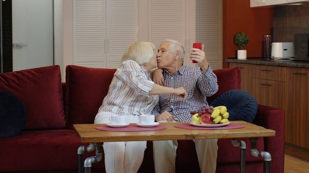 Senioren küssen sich, machen Selfie-Fotos, nehmen zu Hause gemeinsam Videos auf dem Smartphone auf - Foto, Bild