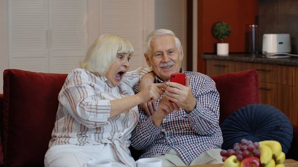 Oudere grootouders die thuis met elkaar praten en mobiele telefoons gebruiken. Internet winkelen - Foto, afbeelding