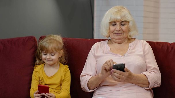 Starsza babcia z wnuczką dziewczynki korzystającą z telefonu komórkowego, grającą w gry w domu - Zdjęcie, obraz