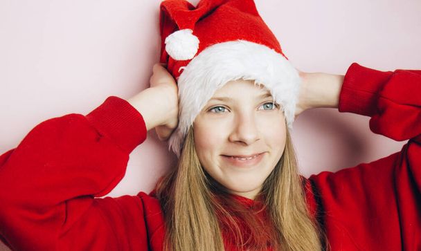 Smiling beautiful girl with long blonde hair in fluffy Santa Claus hat on a pink background. Teenager with bright emotions holding her head with his hands - Zdjęcie, obraz