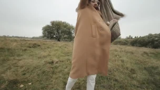 Young brunette in a coat stands against the background of a tree and holds a wide neckerchief waving in the wind - Footage, Video