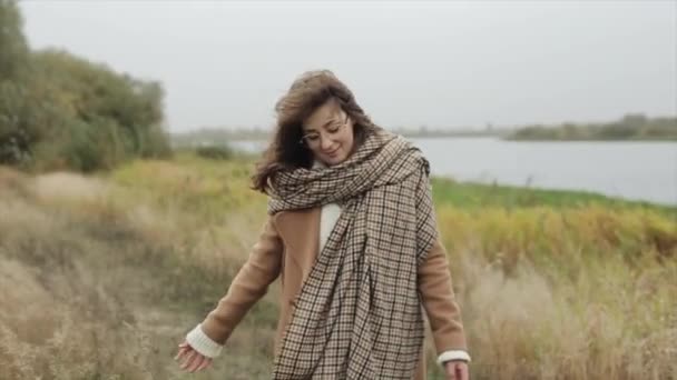Een aantrekkelijke jonge vrouw loopt langs het pad langs de rivier gewikkeld in een sjaal en glimlachend raakt het gras met haar handen. Langzame beweging - Video