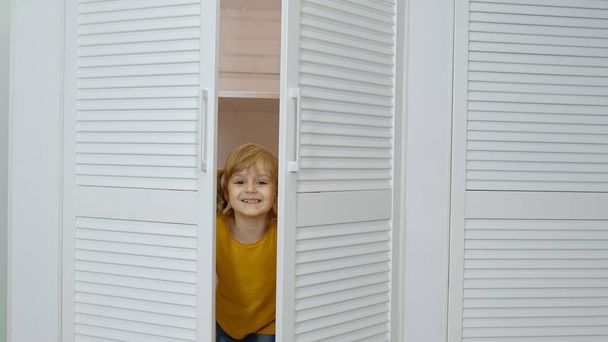 Mała blondynka dziewczynka w dżinsach, pomarańczowa kurtka otwierająca szafę i patrząca w kamerę w domu - Zdjęcie, obraz