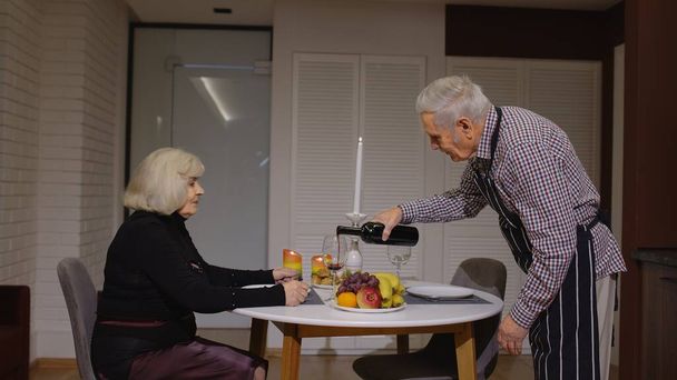 Vanhempi eläkeläispariskunta pitää hauskaa juomalla viiniä ja syömällä aterian aikana romanttinen illallinen keittiössä - Valokuva, kuva