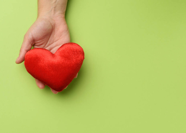 mano femenina sostiene corazón textil rojo, fondo verde. Concepto de amor y donación, de cerca - Foto, imagen