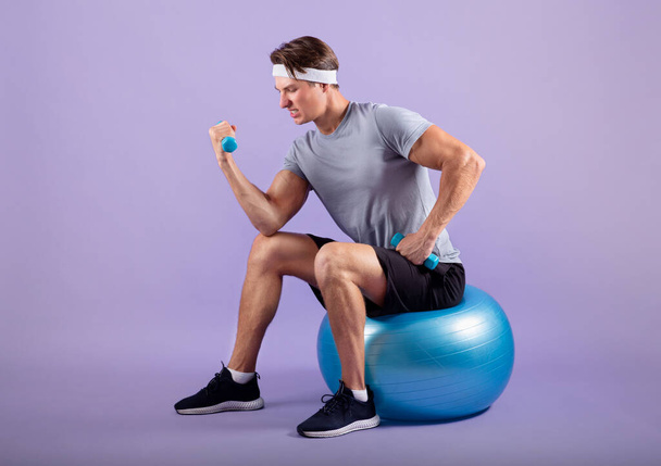 Determinato sportivo millenario che si allena con i manubri mentre siede sulla palla da fitness su sfondo lilla - Foto, immagini