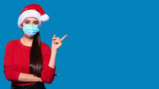 Joven mujer de Santa en la máscara de la cara del sombrero de Navidad señalando dedo índice en el producto sobre fondo aislado azul. - Foto, imagen
