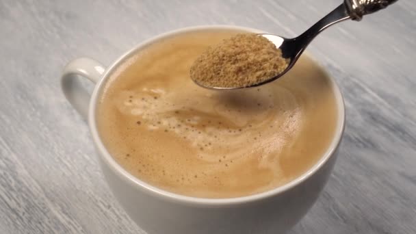 Hand adds cane sugar with a dessert spoon to a white ceramic cup with frothy coffee. Stirring. Close up - Video