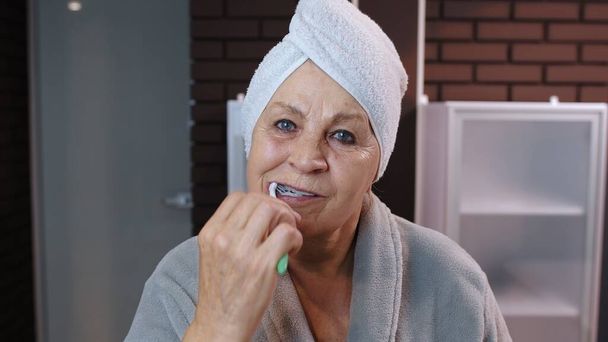 Старша бабуся з зубами для чищення рушників і заглядає в дзеркало після душу вдома
 - Фото, зображення