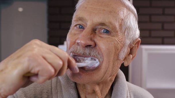 Старший дед в халате, чистит зубы, смотрит в зеркало. Утренняя гигиена в ванной - Фото, изображение