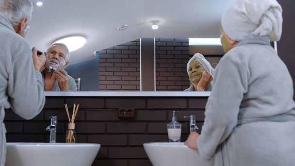 Старшая бабушка надевает маску для лица и дедушка бреется ручным лезвием бритвы в ванной - Фото, изображение