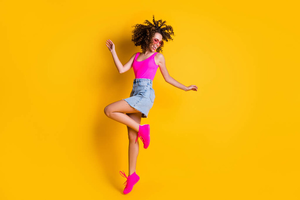 Full body photo of attractive youngster curly lady jump up high students party rejoicing wear sun specs pink tank top denim mini skirt shoes isolated vivid bright yellow color background - Zdjęcie, obraz