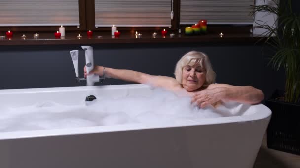 Sexy anciana anciana rubia abuela está tomando un baño espumoso en el baño de lujo con velas - Imágenes, Vídeo